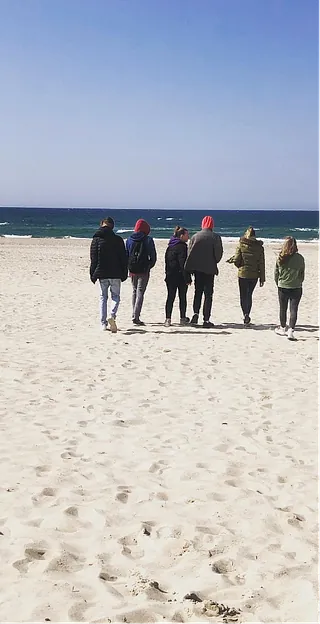 Sechs Personen gehen bei sonnigem Wetter gemeinsam über einen Sandstrand in Richtung Meer.