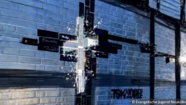 Eine Ziegelwand. Einige Ziegel sind gefärbt, so dass das Jesus Kreuz zu sehen ist. Darüber ist ein weißes Jesus Kreuz als Graffiti an die Wand projiziert.